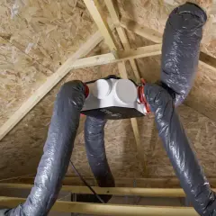 Installation d'une VMC dans un grenier. Le caisson blanc est suspendu, relié à des gaines grises flexibles.