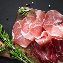 Cienkie plastry prosciutto, w tym szynka parmeńska, ułożone na drewnianej desce z gałązkami rozmarynu i ziarnami pieprzu.
