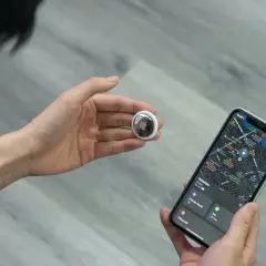 Osoba trzymająca w dłoni lokalizator AirTag i iPhone'a z mapą. AirTag co to? To małe urządzenie do śledzenia zgubionych przedmiotów.