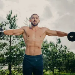 Mężczyzna z szerokimi barkami ćwiczy na świeżym powietrzu, podnosząc hantle.