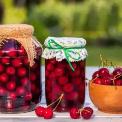 Dwa słoiki domowego kompotu z wiśni, obok miseczka świeżych owoców.