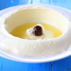 Un plato blanco con labneh que es un queso cremoso, con aceite de oliva y una aceituna negra en el centro, sobre una mesa azul.