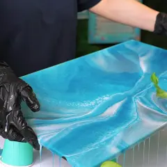 Mani guantate controllano un'opera d'arte in resina epossidica blu e bianca, simile al mare. Il tempo di indurimento della resina epossidica è cruciale per questo effetto.