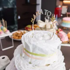 Una torta di pannolini decorata con un topper "Mama to Be" per un baby shower. Sul tavolo anche cupcakes colorati e dolcetti.