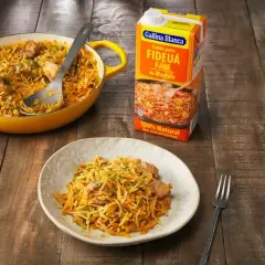 Un plato de fideos a la cazuela servido en una cazuela amarilla y en un plato blanco, junto a un cartón de caldo para fideuá.