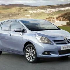 En ljusblå Toyota Verso står parkerad på en väg med en lantlig by i bakgrunden. Kanske är det ett problem med bilen som gör att den står stilla.