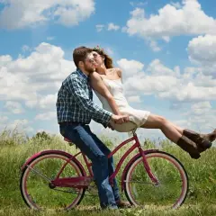 Coppia su bici rosa, lei seduta davanti a lui, baci appassionati sotto un cielo azzurro. Ecco perchè ci si innamora.