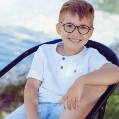 Uśmiechnięty chłopiec w okularach, 7 latek, siedzi na krześle przy wodzie.
