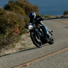 Motociclista inclinado en una curva, demostrando el **efecto giroscópico** de las ruedas al girar. El mar y la vegetación bordean la carretera.