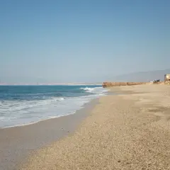 La almadraba de Monteleva, una torre blanca se alza en la playa, con el mar azul y las montañas al fondo.