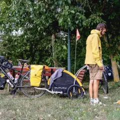Un homme en jaune se tient près d'un vélo chargé, avec une remorque et un jeune chien joueur. Un chien Canada prêt pour l'aventure.