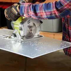 A person in a plaid shirt and work gloves uses a circular saw to cut a sheet of plexiglass, demonstrating how to cut plexiglass cleanly.
