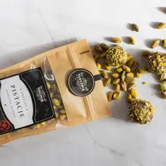 Opakowanie pistacji i trzy praliny z pistacjami rozsypane na marmurowym blacie. Warto sprawdzić pistacje kcal przed spożyciem.