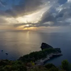 Atardecer en el mirador de Sa Foradada, con rayos de sol filtrándose entre nubes y dos barcos en el mar.
