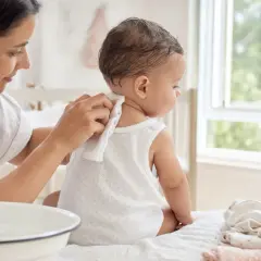 Madre limpia la nuca de su bebé para prevenir la sudamina. El bebé, sentado, mira hacia la ventana.