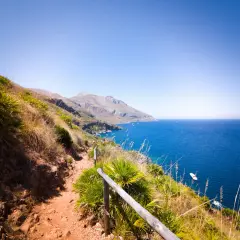 A scenic hiking trail winds through the rugged landscape of Lo Zingaro Nature Reserve, overlooking the brilliant blue Mediterranean Sea dotted with boats.