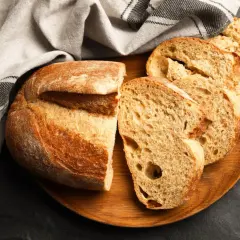 Krojony bochenek chleba i kromki na drewnianej desce, obok lnianej ściereczki.
