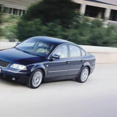 Ciemnoniebieski Volkswagen Passat B5 jedzie po moście, za nim rozmyte drzewa i budynek.