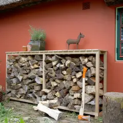 Praktische brennholz lagern ideen: Holzstapel in einem Holzregal vor einer roten Wand, daneben ein Baumstumpf und eine Axt.