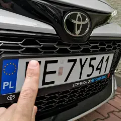 Palec wskazuje na tablicę rejestracyjną samochodu, która wygląda na zgubioną.