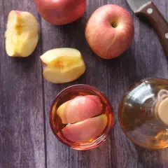Jabłka i szklanka z kawałkami jabłek, obok butelka z płynem, który może być domowym **octem jabłkowym**.