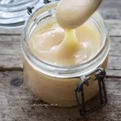 Un cucchiaio preleva il cremoso latte condensato da un barattolo di vetro su un tavolo di legno rustico.