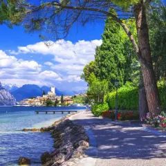 Promenade ensoleillée au bord du lac de Garde, avec des montagnes majestueuses en arrière-plan et un village pittoresque.