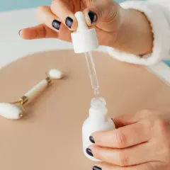 Dłoń z ciemnym lakierem do paznokci trzyma pipetkę z serum, obok jadeitowy roller. Pamiętaj, z czym nie łączyć niacynamidu.