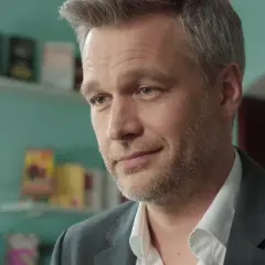 Michał Żebrowski w jednym z filmów, z siwiejącymi włosami i zarostem, w białej koszuli i szarym garniturze.