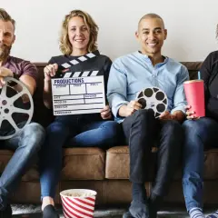 Cztery osoby siedzą na kanapie, trzymając rekwizyty filmowe: klaps, rolki taśmy filmowej, popcorn i napój. Różne rodzaje filmów i ich tworzenie.