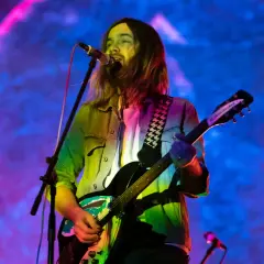 Kevin Parker z Tame Impala gra na gitarze podczas koncertu, w tle kolorowe światła.