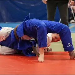 Två judokas i blå och vita dräkter kämpar på en röd matta. En av dem, med ett leende, utför en rörelse inspirerad av Jigoro Kano.