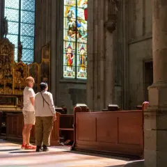 Zwei Besucher bestaunen die bunten Glasfenster im Kölner Dom. Sonnenlicht fällt durch die Fenster und malt bunte Muster auf den Boden.