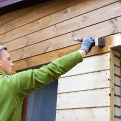 Mężczyzna w zielonej bluzie maluje pędzlem drewnianą elewację domu, chroniąc ją przed warunkami atmosferycznymi.