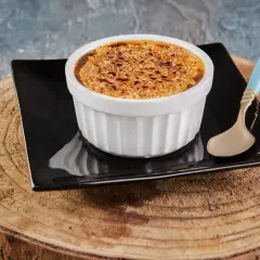 Crème brûlée avec une croûte caramélisée dorée, servie dans un ramequin blanc sur une assiette noire, avec une cuillère à la touche champignon.