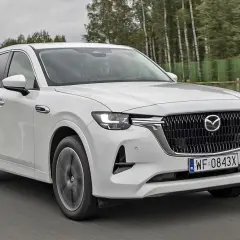 Biała Mazda CX-60 jedzie po drodze, otoczona drzewami. Sprawdź, jaka jest mazda cx 60 cena i wyposażenie.