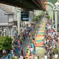 Bunte Farben auf dem Boden und eine Schwebebahn über der belebten Straße im Wuppertal Zentrum. Menschenmassen säumen die bunte Gasse.