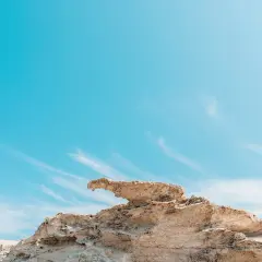 Roca erosionada con forma de ave en una playa de arcos, bajo un cielo azul intenso con nubes finas.