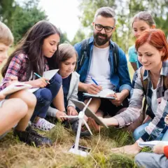 Grupa dzieci i dwoje dorosłych siedzi na trawie, obserwując model turbiny wiatrowej i notując w zeszytach.