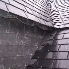 Détail d'une toiture ardoise, avec des ardoises sombres et brillantes, fixées par des crochets métalliques.