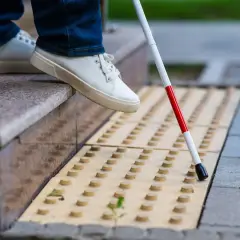 Osoba w białych butach i jeansach używa białej laski z czerwonym paskiem na żółtej nawierzchni z wypustkami.