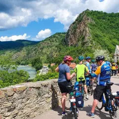 A group of cyclists on a Danube bike tour pauses by a stone wall, with a river and lush green hills in the background.