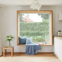 Gemütliche Fensterbank mit Kissen und Decke, ein Buch liegt offen. Das große Fenster ohne Gardinen gibt den Blick auf einen üppigen Garten frei.