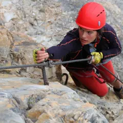 Młoda kobieta w kasku wspina się po skale, używając stalowej liny. To przygoda na via ferrata Czechy.