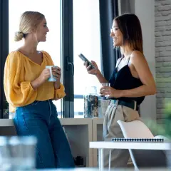 Dwie kobiety rozmawiają przy oknie, jedna z kubkiem, druga z telefonem. To przykład, jak wygląda **small talk co to** jest.