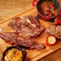 Un plateau en bois présente une entrecôte barbecue juteuse, des morceaux de viande braisée, des sauces et des légumes.