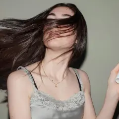 Mujer con cabello largo y oscuro moviéndose, sosteniendo un frasco de **aceite para el pelo**.