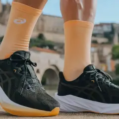 Biegacz w pomarańczowych skarpetkach z logo firmy Asics i ciemnych butach sportowych na tle mostu i zabudowań.