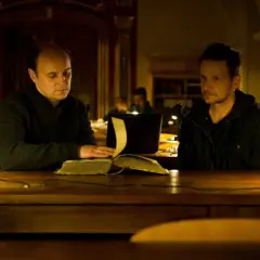 Dwóch mężczyzn przy stole w bibliotece, jeden czyta starą księgę. Scena z filmu "Sługi Boże".