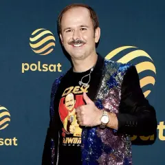 Sławomir Zapała w błyszczącej marynarce i koszulce z własnym wizerunkiem, pokazuje kciuk w górę.